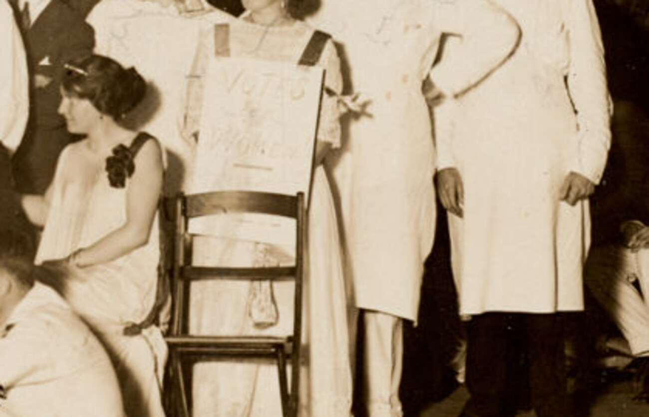 The same photograph, enlarged: a hand-lettered sandwich board reading VOTES FOR WOMEN, worn by one of the party.