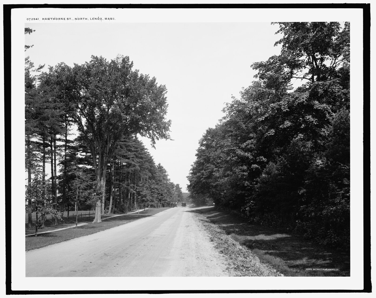 An empty macadam road under tall trees, a post-and-rail fence on the left, a carriage far off in the distance