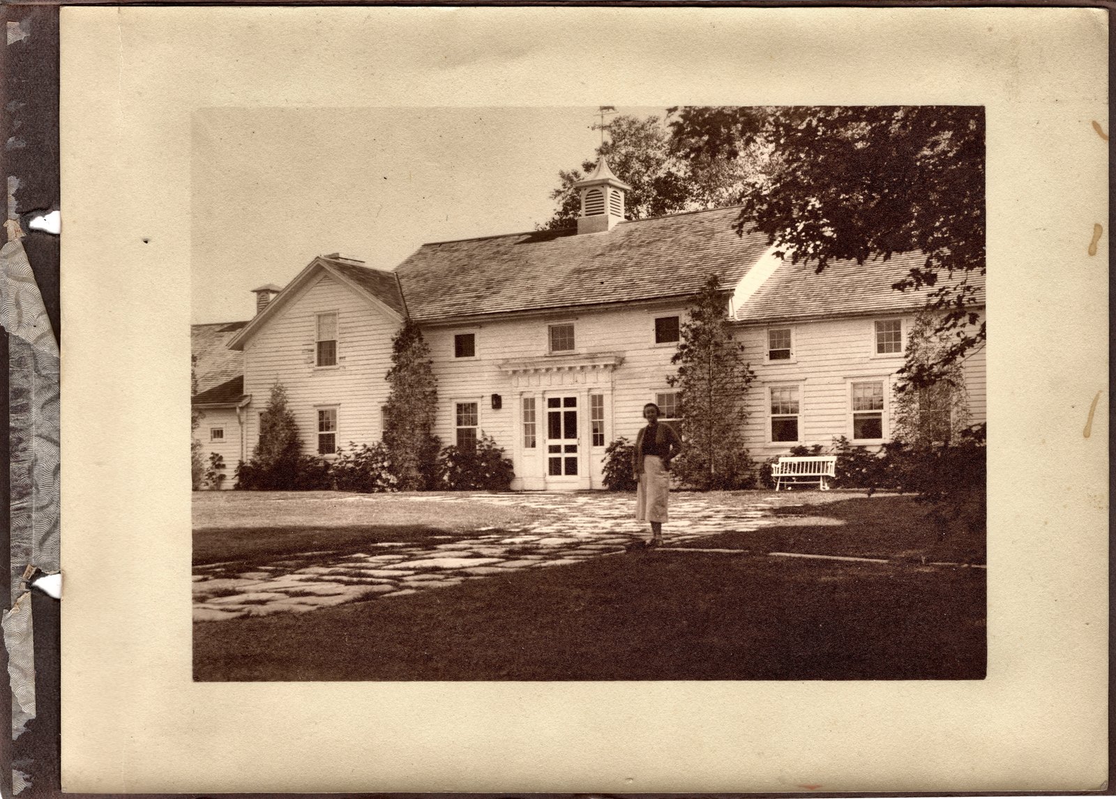 The farm house at Orleton Farm: a long white clapboard house with a cupola, a figure standing on the flagstone walk in front of it