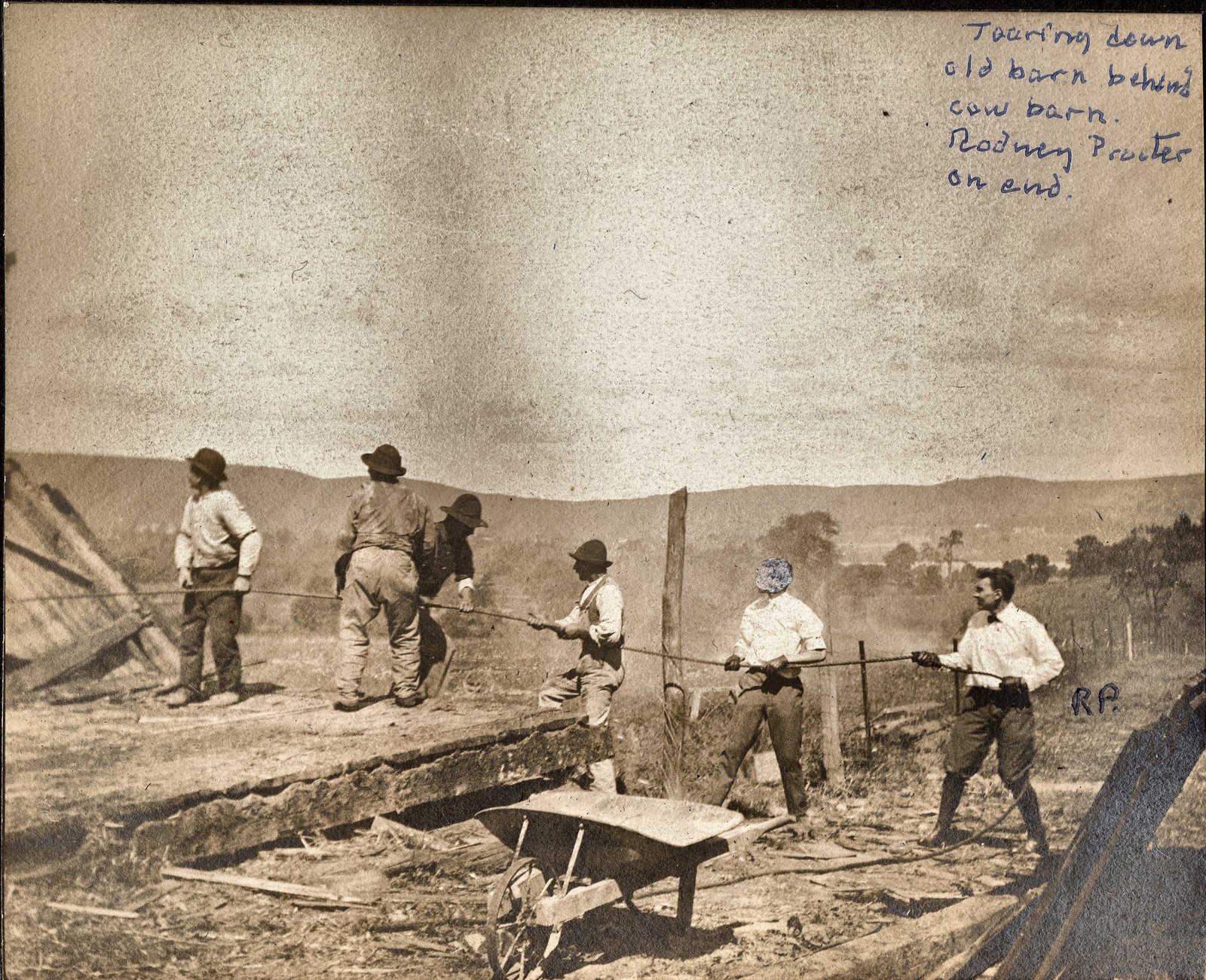 Tearing down old barn behind cow barn. Rodney Procter on end. The figure at the right of the frame is initialed RP on the print.