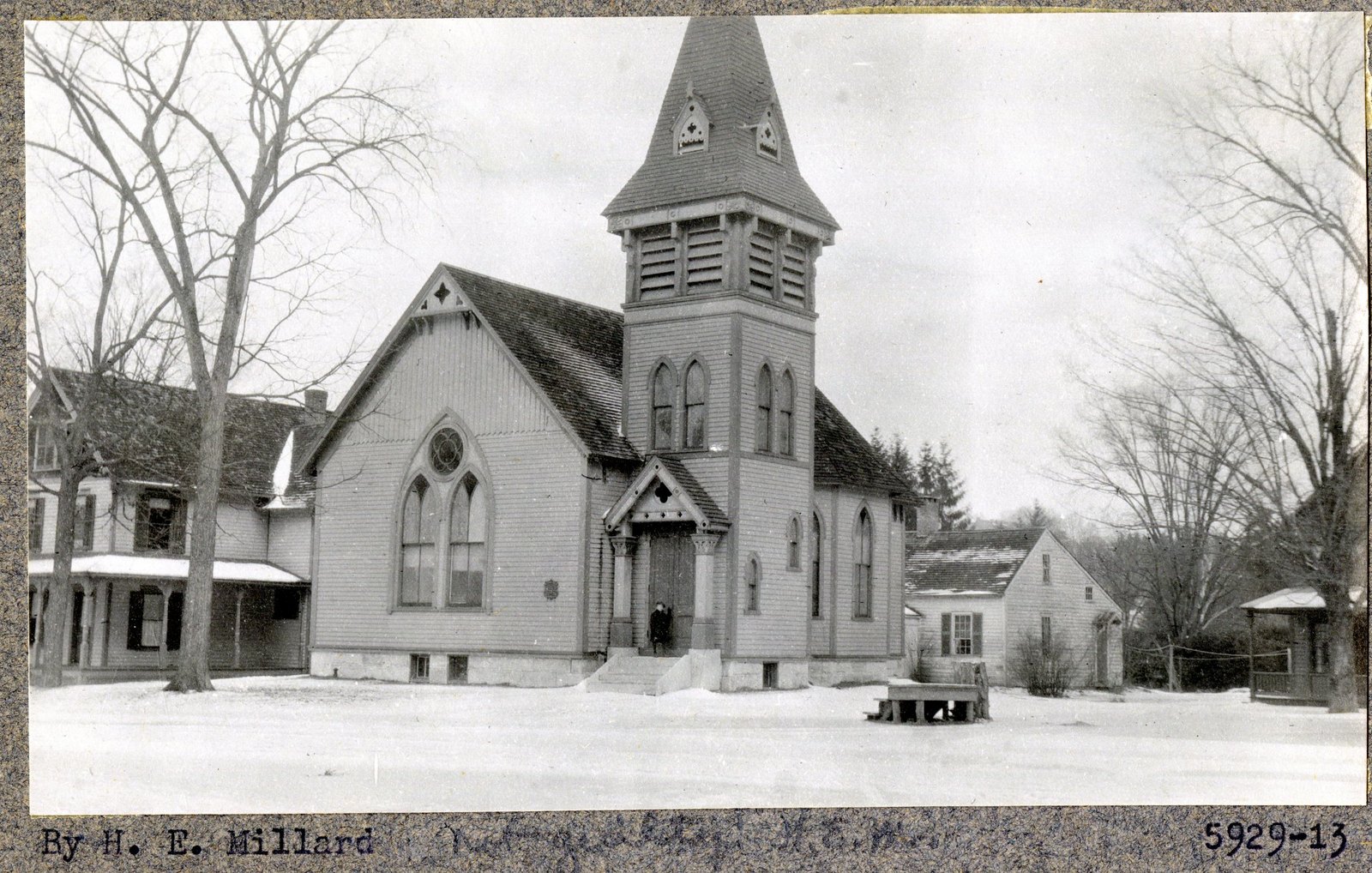 Print 5929-13, credited in type “By H. E. Millard,” with a pencil note beside it in a lighter hand. A clapboard church with a louvred belfry and a gabled porch, a figure standing in the doorway.