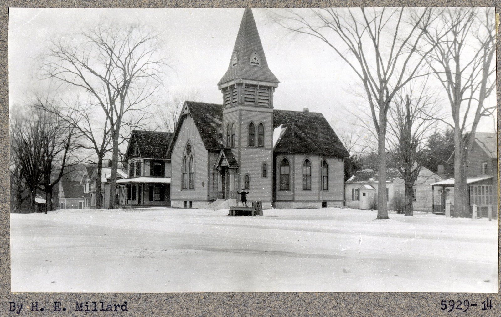 Print 5929-14, credited “By H. E. Millard.” The same church from further back, snow across the whole foreground, a figure on the porch platform.